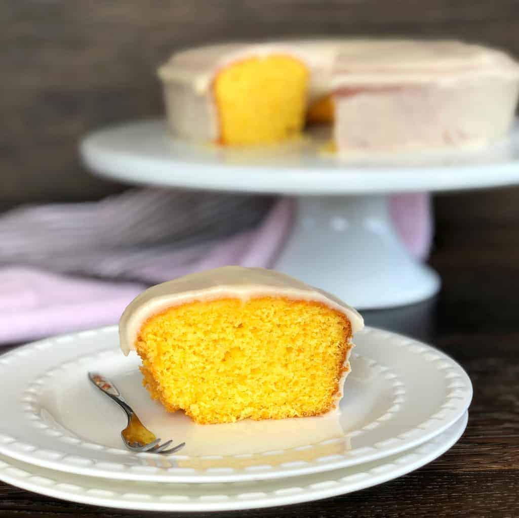 Slice of Magical Orange Cake on a white plate with the rest of the cake on a white cake stand in the background.