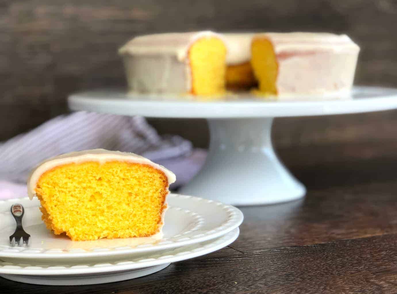 Slice of Magical Orange Cake with rest of cake in background on a white cake stand