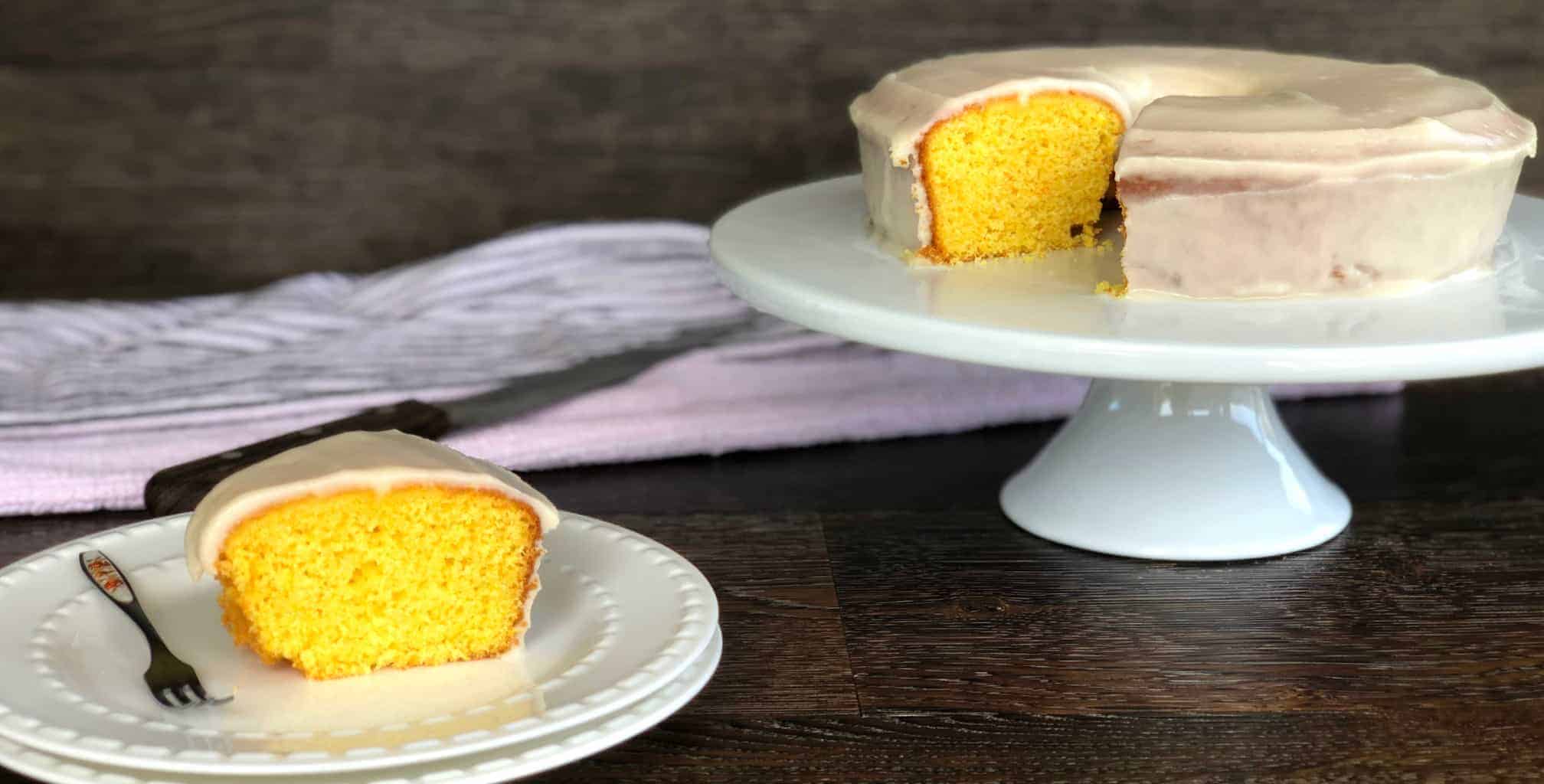 Slice of Magical Orange Cake on a white plate. With the rest of the cake on a white cake stand in the back.