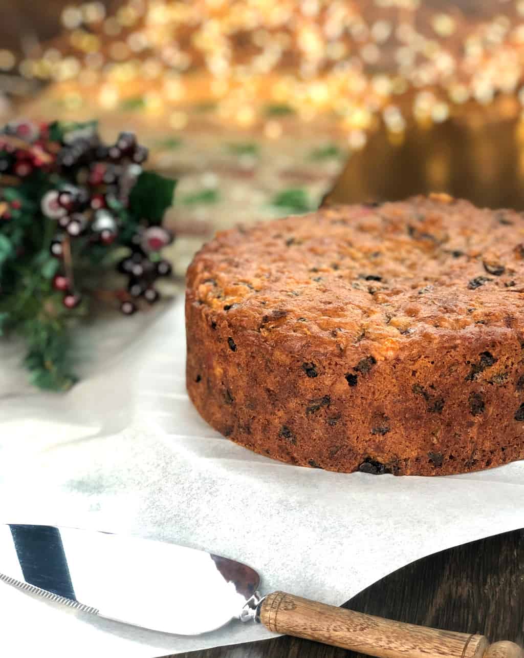 Christmas Cake next to a serving slice with Christmas Holly and Berries in the background.