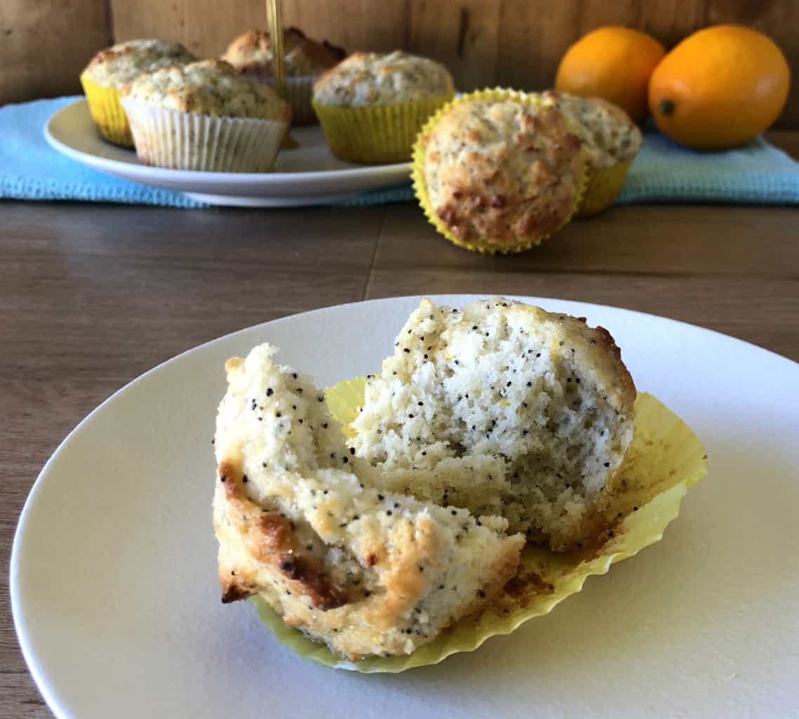 Lemon and Poppyseed muffin torn open to show the soft fluffy inside with other muffins in the background and fresh lemons