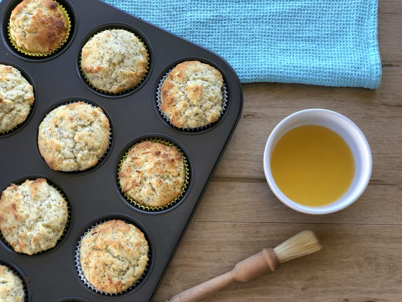 Freshly baked muffins in a muffin tin with a sugar lemon glaze prepared and a pastry brush