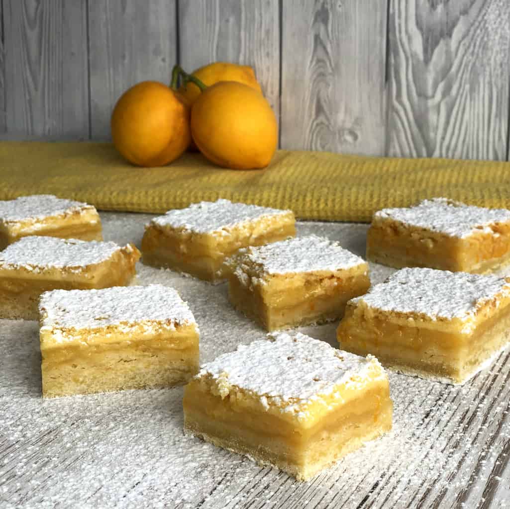 Individual Slices of Lemon Squares baking on a white background dusted in icing sugar and lemons in the background on a yellow cloth
