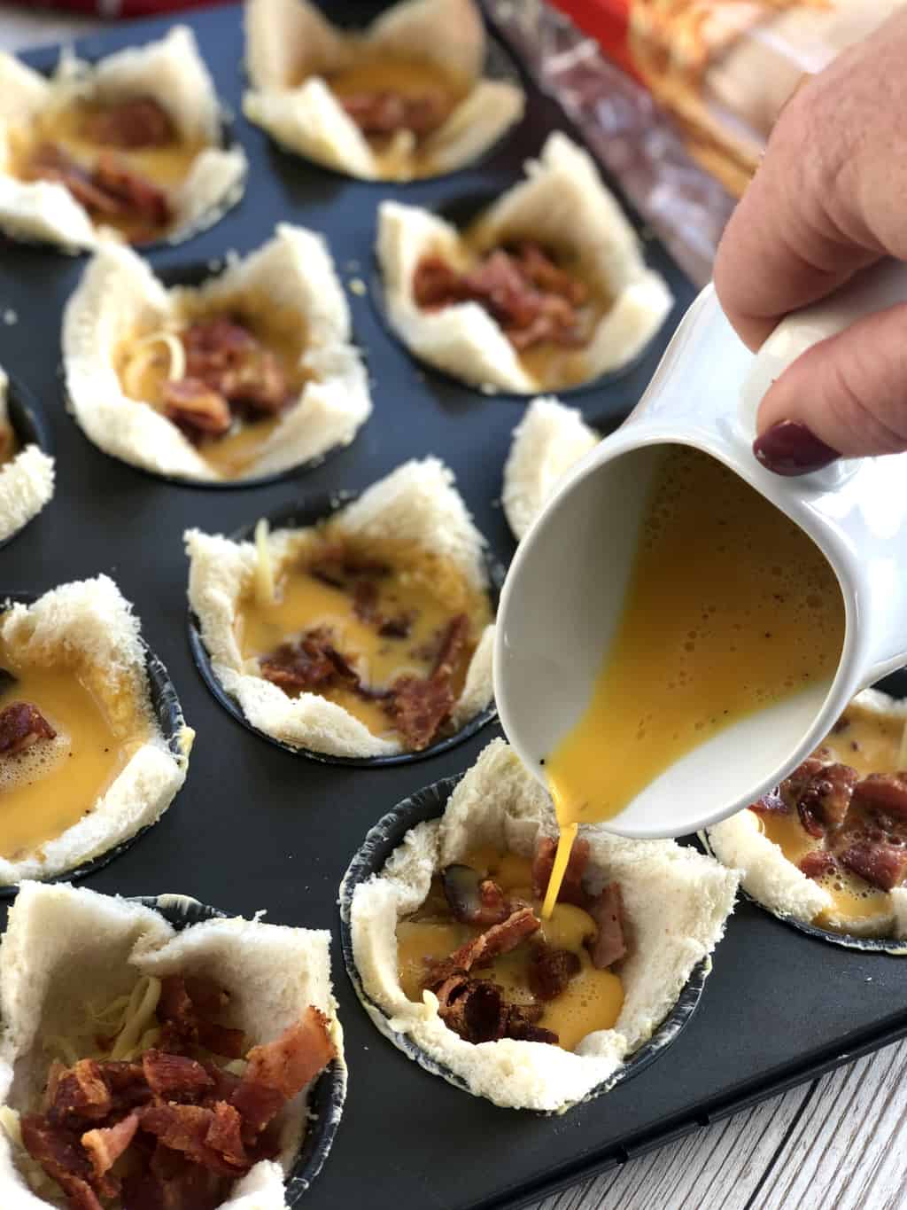 a white jug is pouring egg mixture into the bacon and cheese filled bread cases