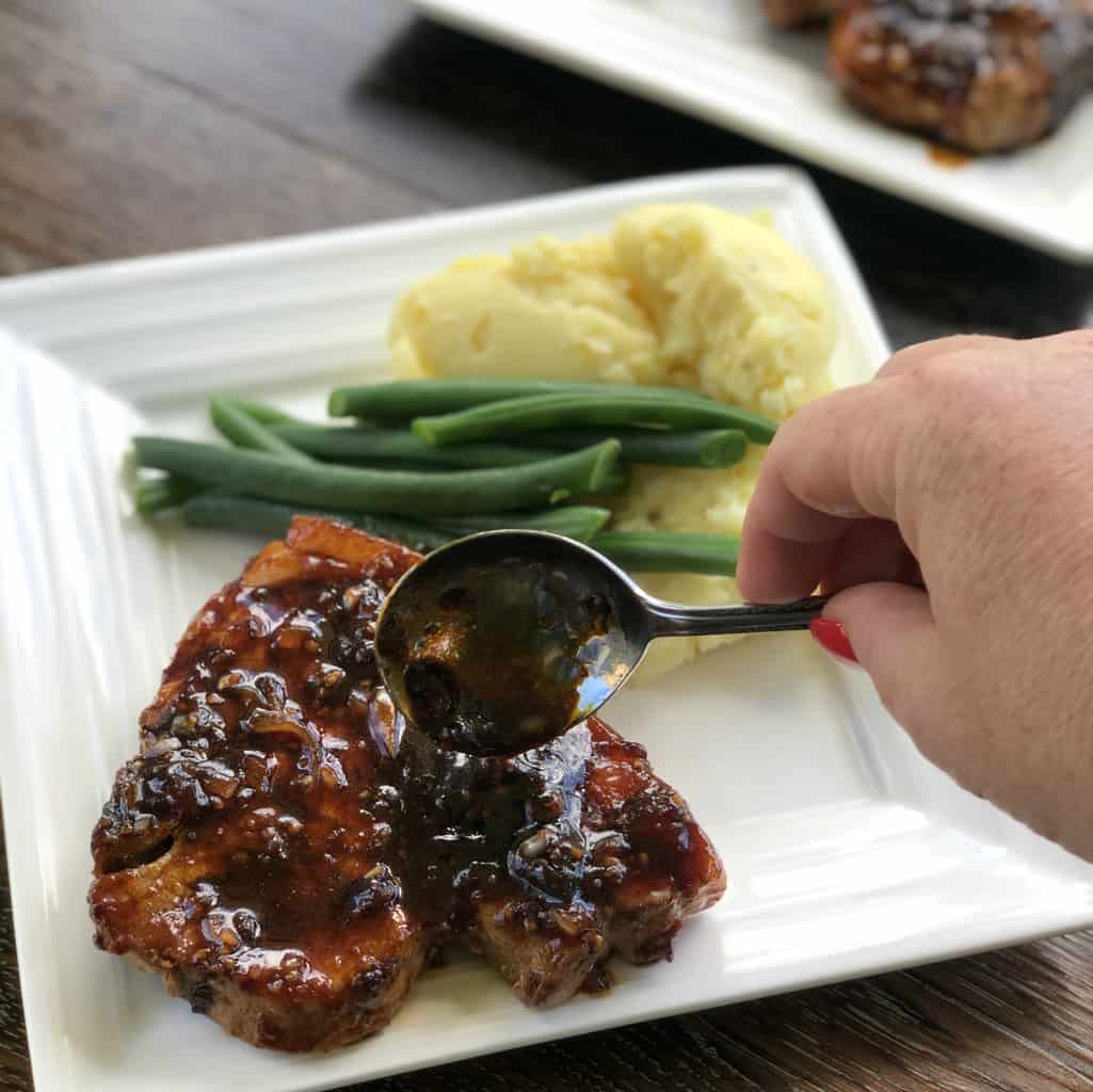 Spooning an apricot and soy glaze over a pork chop on a white plate with side dishes