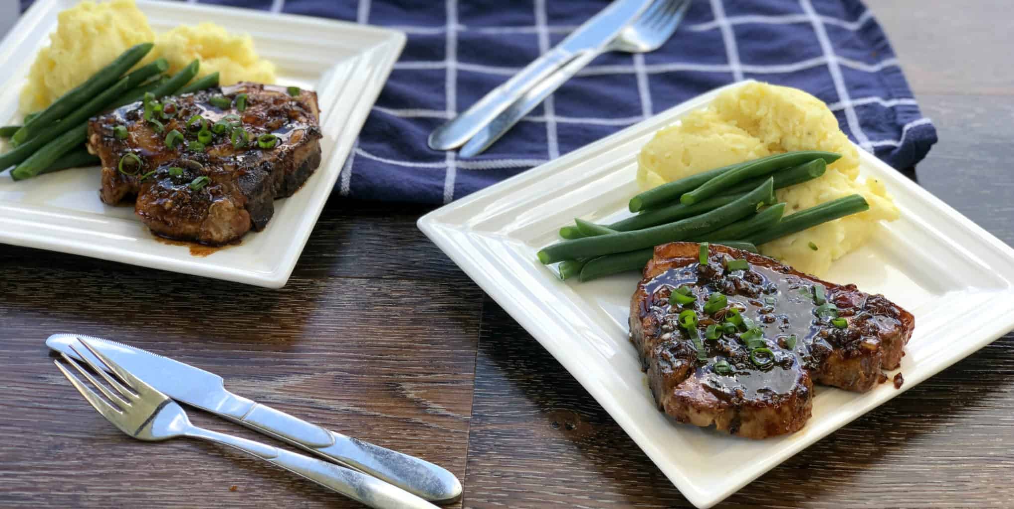 Two white plates prepared with pork chops, glazed, mashed potatoes and green beans 