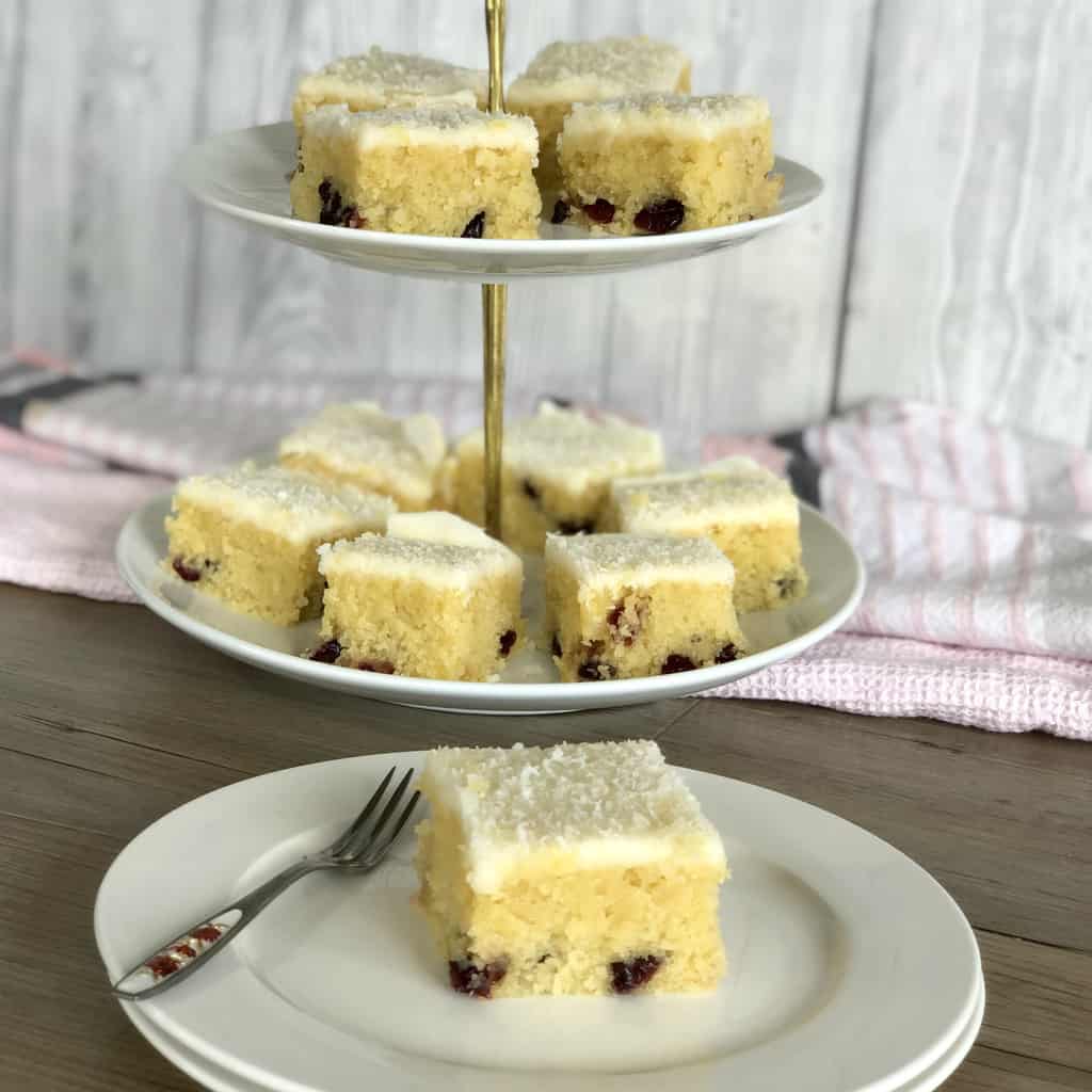 White tiered plate with slices of white coconut slice and cranberries and a white plate with a piece of slice and a decorative fork