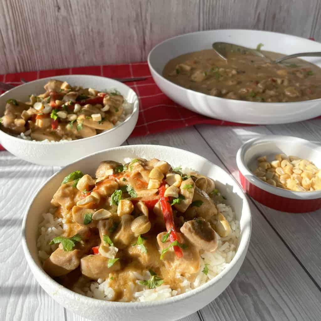 Two bowls of Slow Cooker Satay Chicken in white bowls on rice, with a large bowl of the recipe in the background, garnished with coriander and cashews