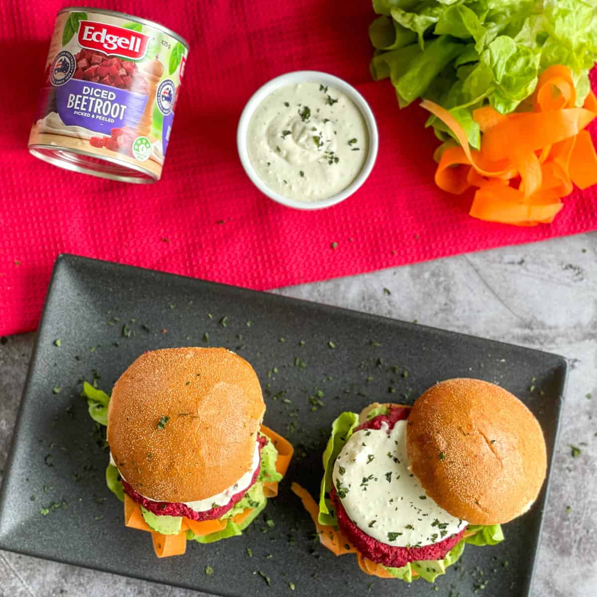 Overhead photograph of a red cloth with two vegetarian burgers and a bowl of whipped feta