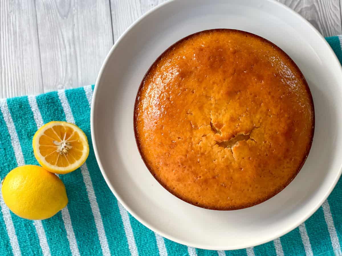 Overhead photograph of lemon syrup cake on a white platter and fresh lemons on a green cloth