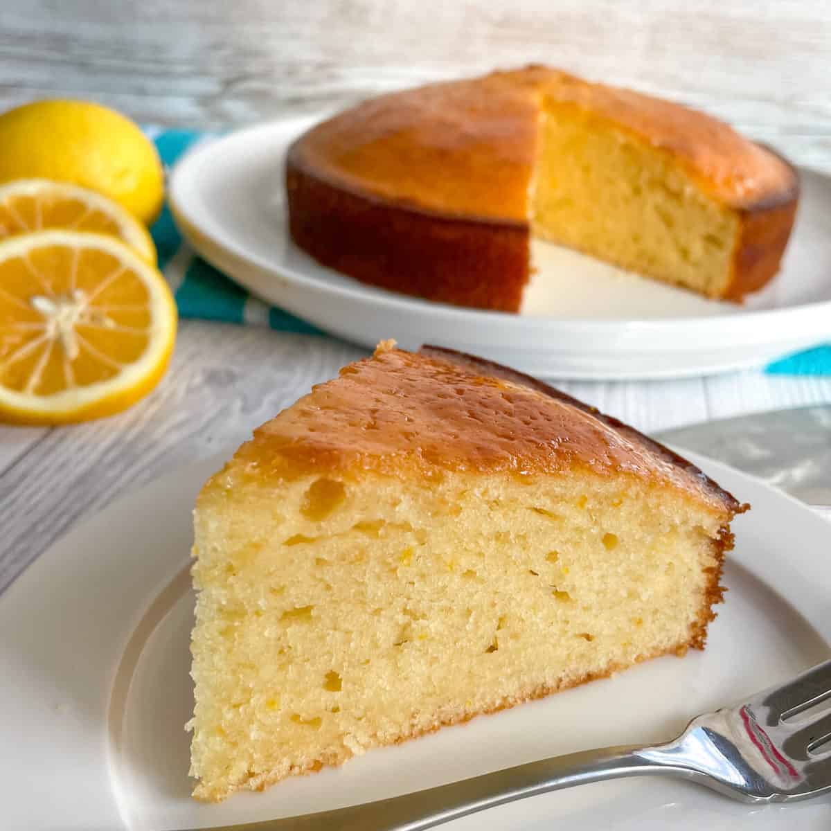 Slice of lemon syrup cake on a white plate, with the rest of the cake in the background