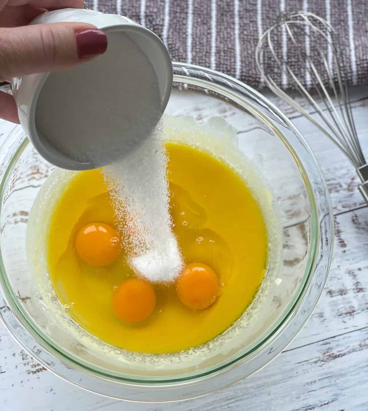 Pouring caster sugar into a glass bowl containing melted white chocolate, butter and eggs 