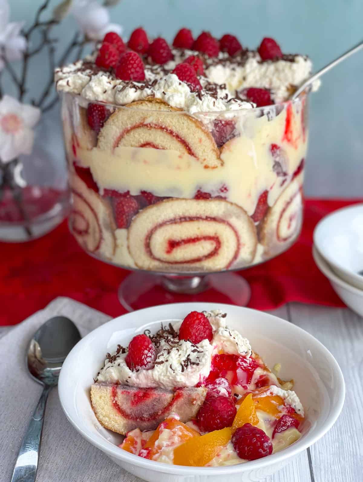 A bowl of trifle served with peaches, custard, sponge roll, jelly, strawberries and cream