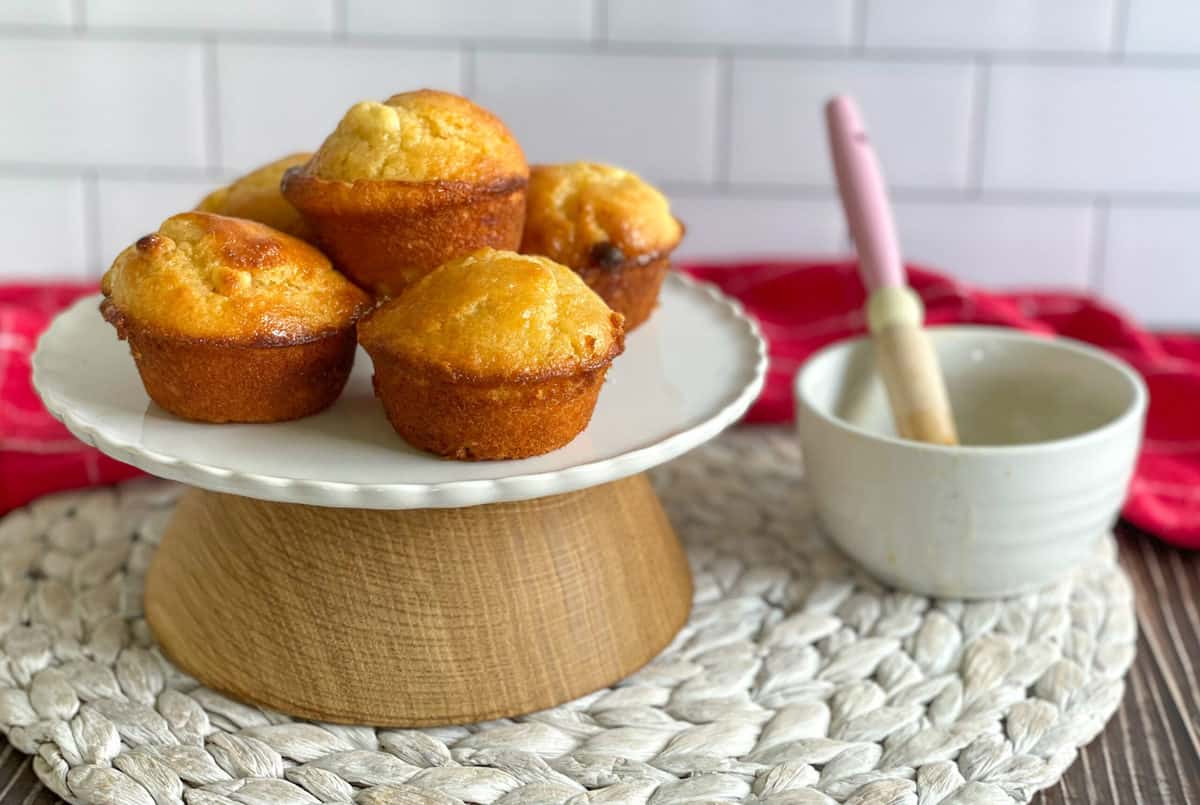 Golden passionfruit muffins on a white and wooden platter with a passionfruit syrup and pastry brush nearby