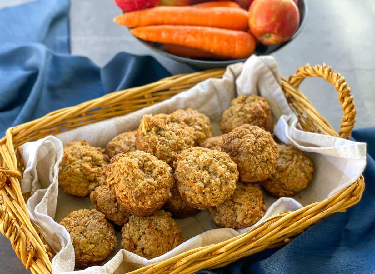 Wicker basket with a tea towel and loaded with apple and carrot muffins and a bowl of apples and carrots on a blue backgroud