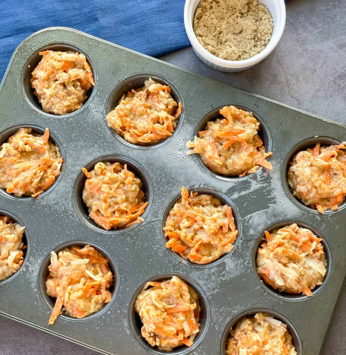 Apple and carrot muffins in a muffin tray before baking with a bowl of sugar for sprinkling 