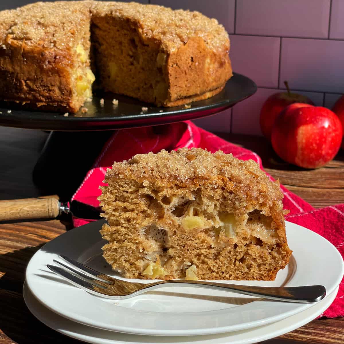 A slice of Apple Crunch Cake with a whole cake and fresh apples in the background