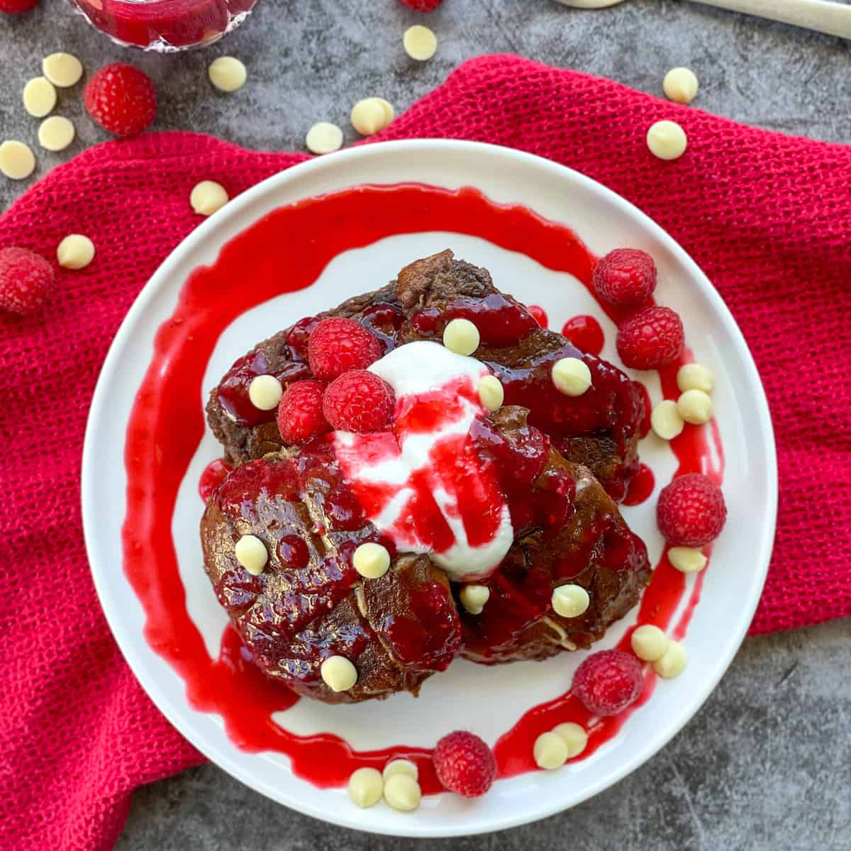 White plate with raspberry coulis and hot cross bun French toast with Greek yoghurt on a red cloth against a concrete background 