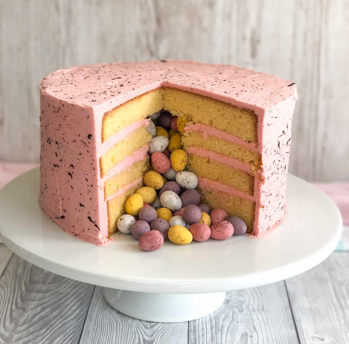 A large 4 layer cake with pink buttercream and easter eggs falling from the centre of the cake on a white background
