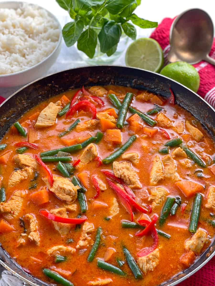 A pan full of chicken and kumara red curry with vegetables and limes, basil and rice in the background