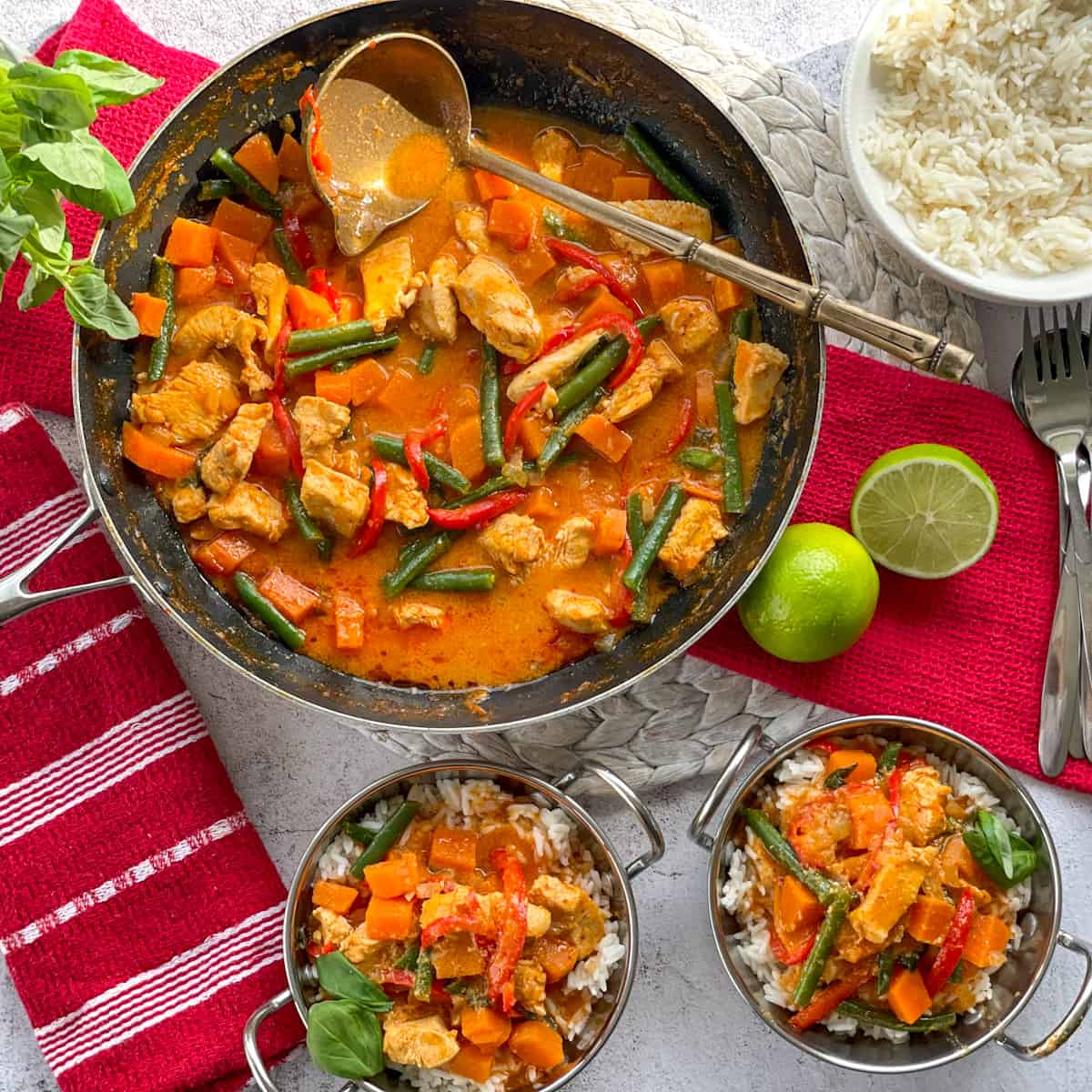 two bowls of red curry with rice, and a pan full of curry with limes, basil and rice to serve