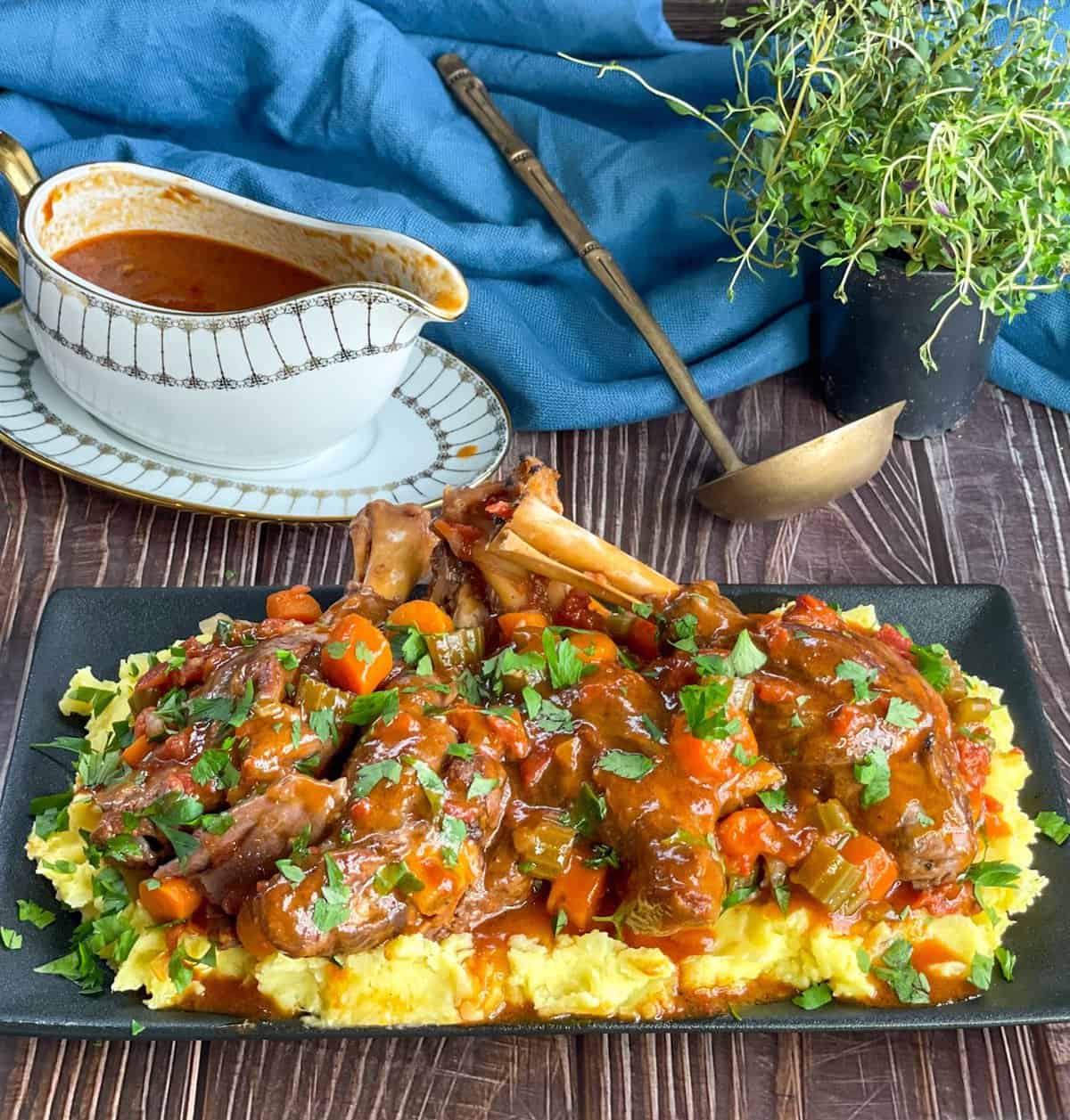 Black platter with mashed potato and lamb shanks and red wine sauce in a gravy boat 