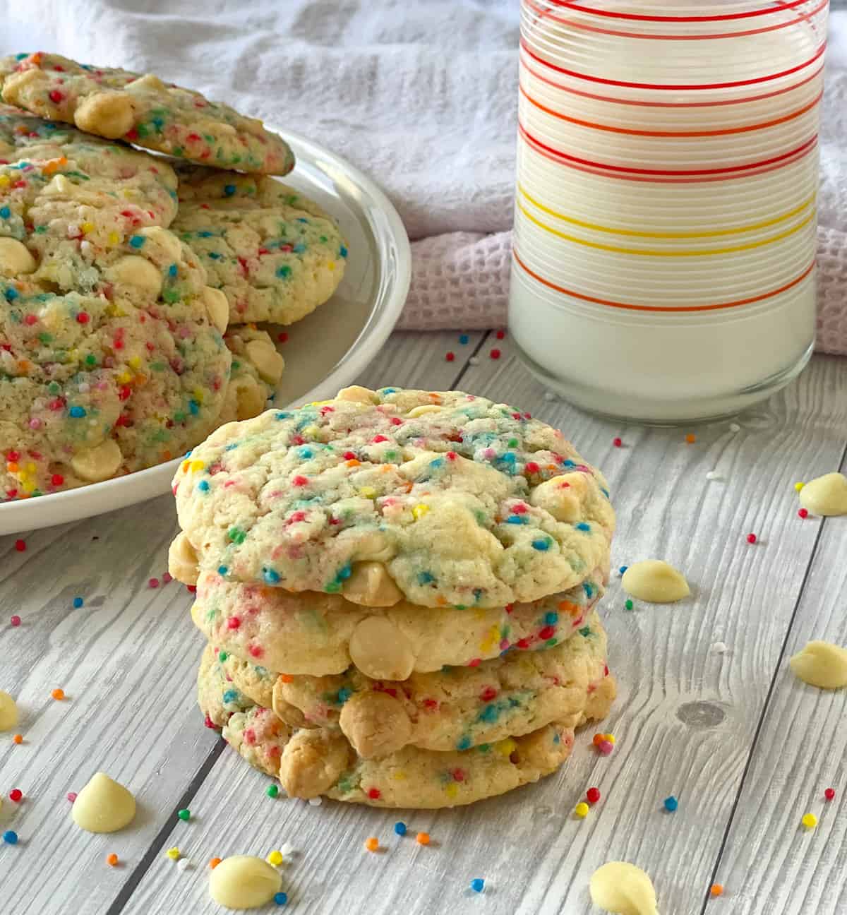A stack of Cake Mix White Chocolate Sprinkle Cookies with a glass of milk and a plate of cookies in the background