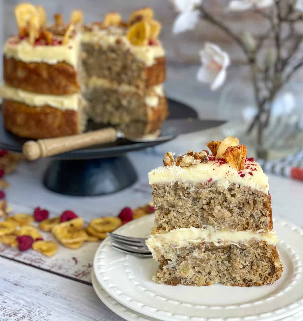 Slice of Hummingbird cake with two layers and cream cheese frosting. The remainder of the cake is on a black cake stand in the background.