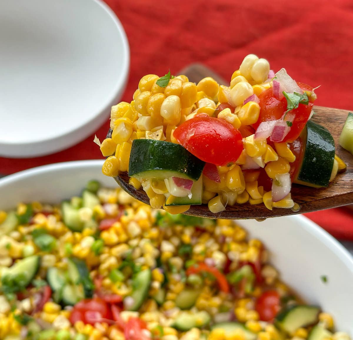 A wooden spoon holding a scoop of sweetcorn salad with tomato, cucumber and red onion