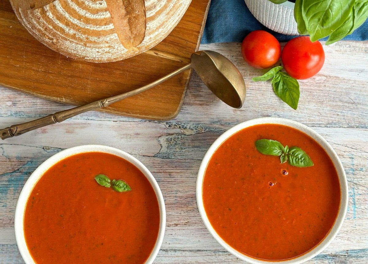 Slow Cooker Roasted Tomato and Basil Soup in two white bowls and a loaf of bread and fresh tomatoes and basil