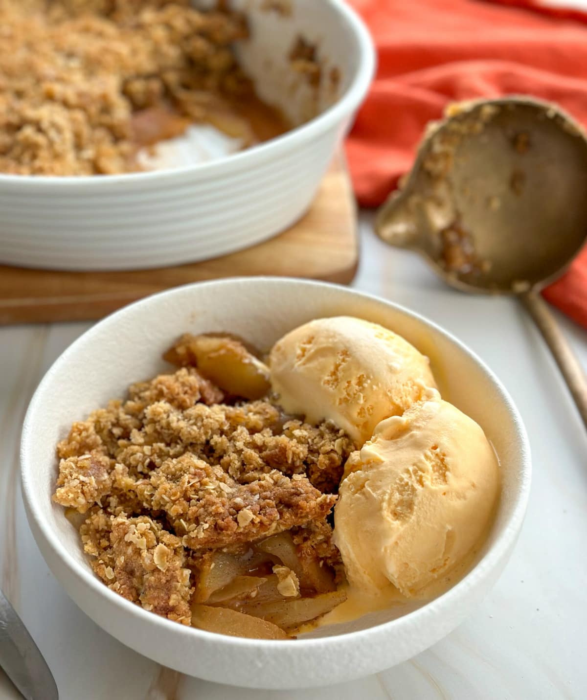 A serving of apple oat crumble crisp, served in a white bowl with two scoops of vanilla ice cream