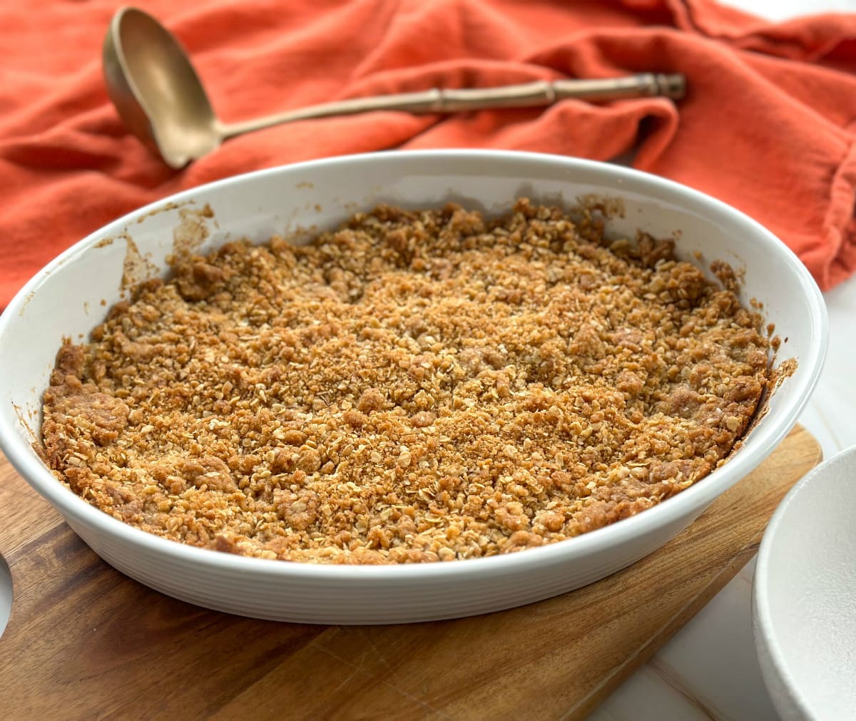 Apple Oat Crumble Crisp in a white dish with an orange cloth and a brass ladle