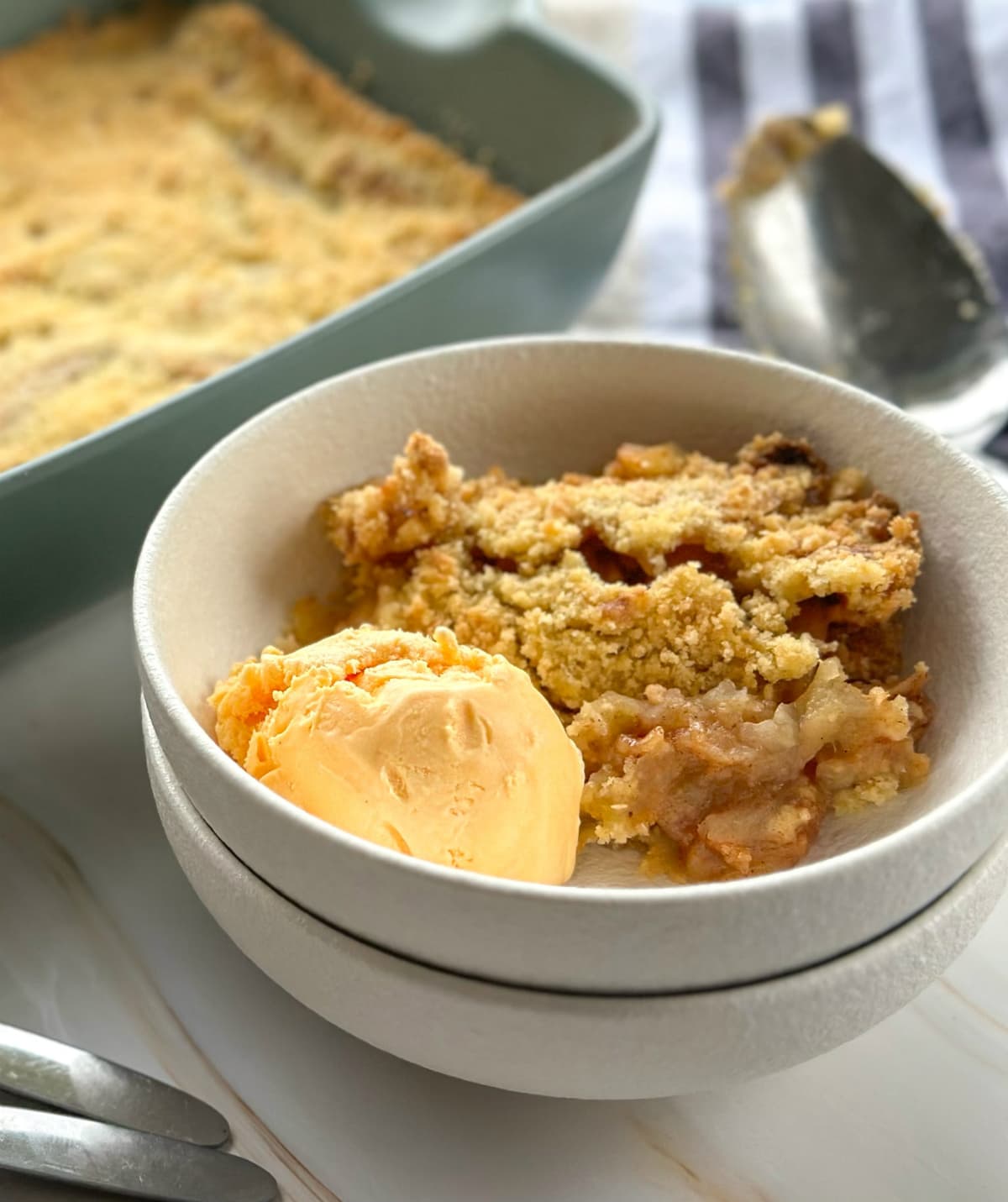 Serving of apple pie with french vanilla ice cream in a white bowl with a dish of apple crumble in the background