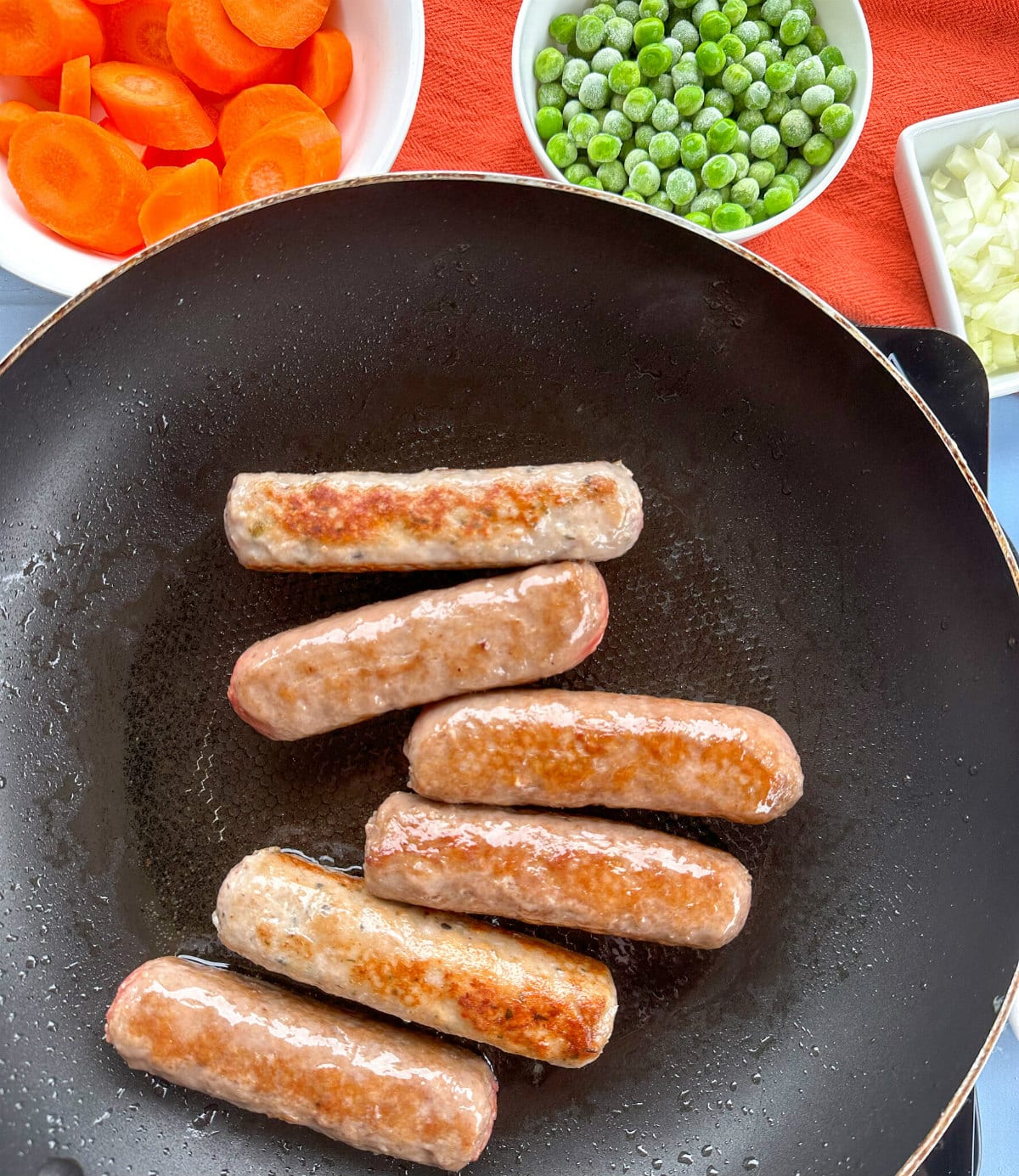 Frying six sausages in a frying pan with bowls of carrots, peas and onions waiting to go in