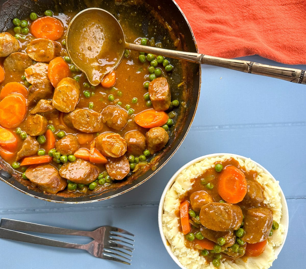 A frying pan full of curried sausages with a bowl of mashed potato and sausages in the forefront