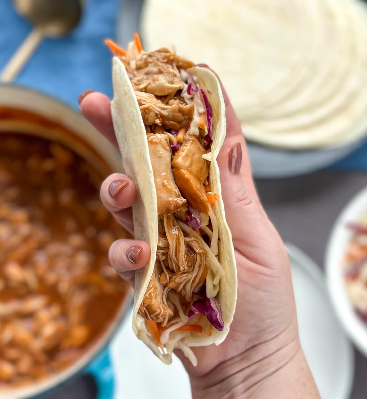 A hand holding a soft taco with coleslaw and BBQ pulled chicken as the filling