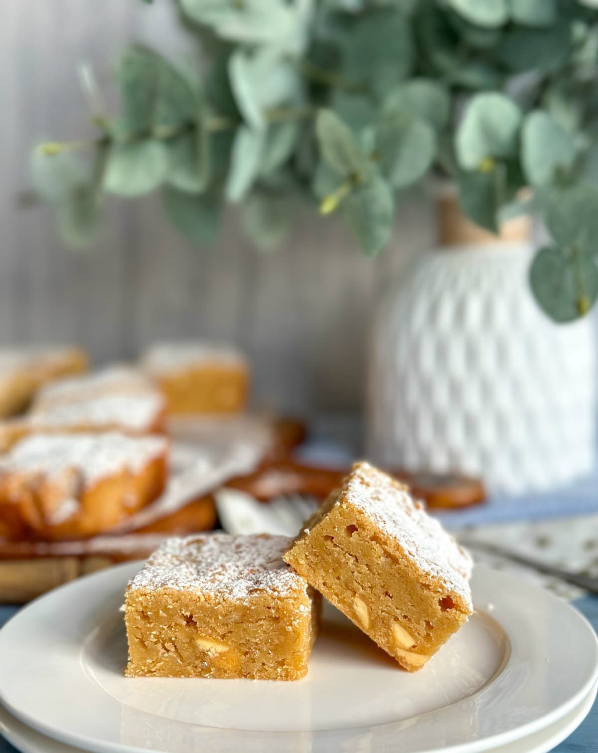 Two slices of blondie on a white plate with baking and floral arrangement in the background 