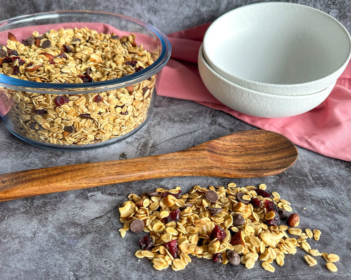 Glass bowl of toasted muesli, a wooden spoon and some muesli scattered in the table with two white dessert bowls