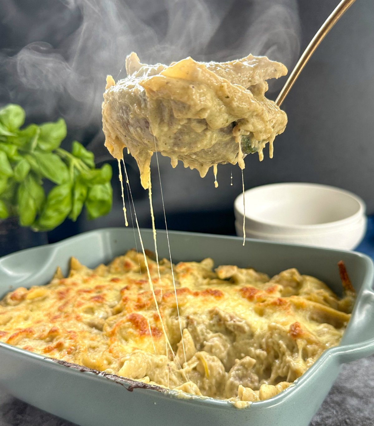 Slow Cooker Creamy Chicken Pesto Pasta Bake in a green baking dish, with mozzarella strings and steam coming off the ladle of pasta