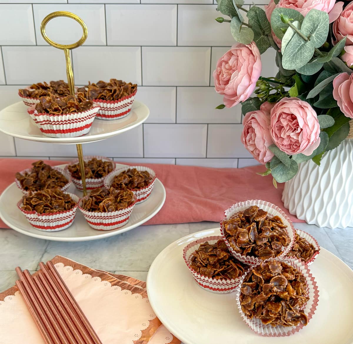 Party table set with chocolate cornflake clusters and a pink background, on a white layered cake stand. 