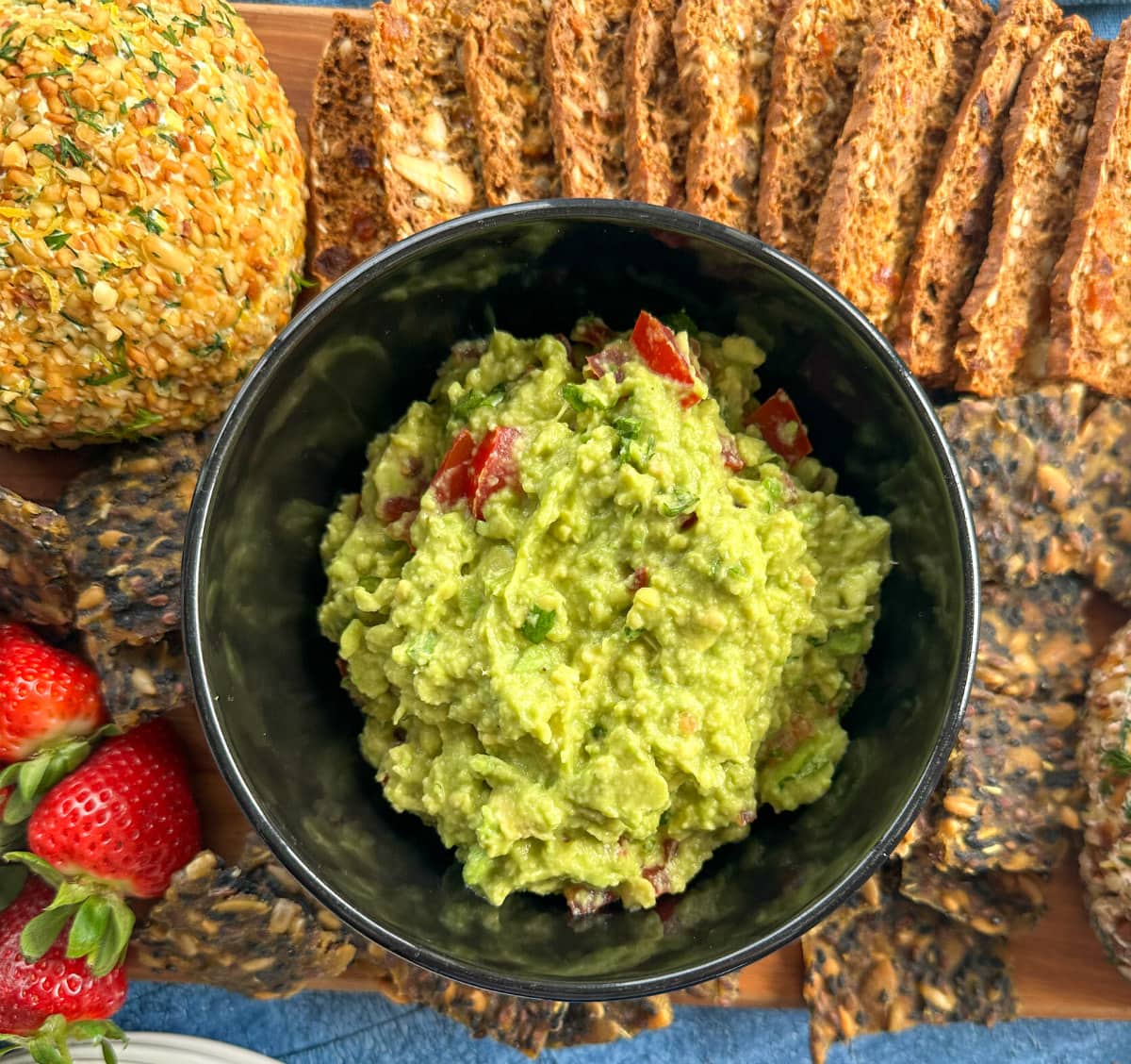The Best Guacamole in a black bowl on a cheeseplatter with crackers and cheeseballs and strawberries