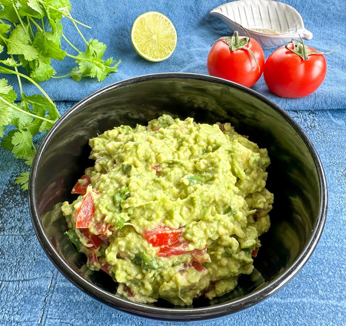 Creamy green guacamole in a black bowl with tomatoes, lime and coriander
