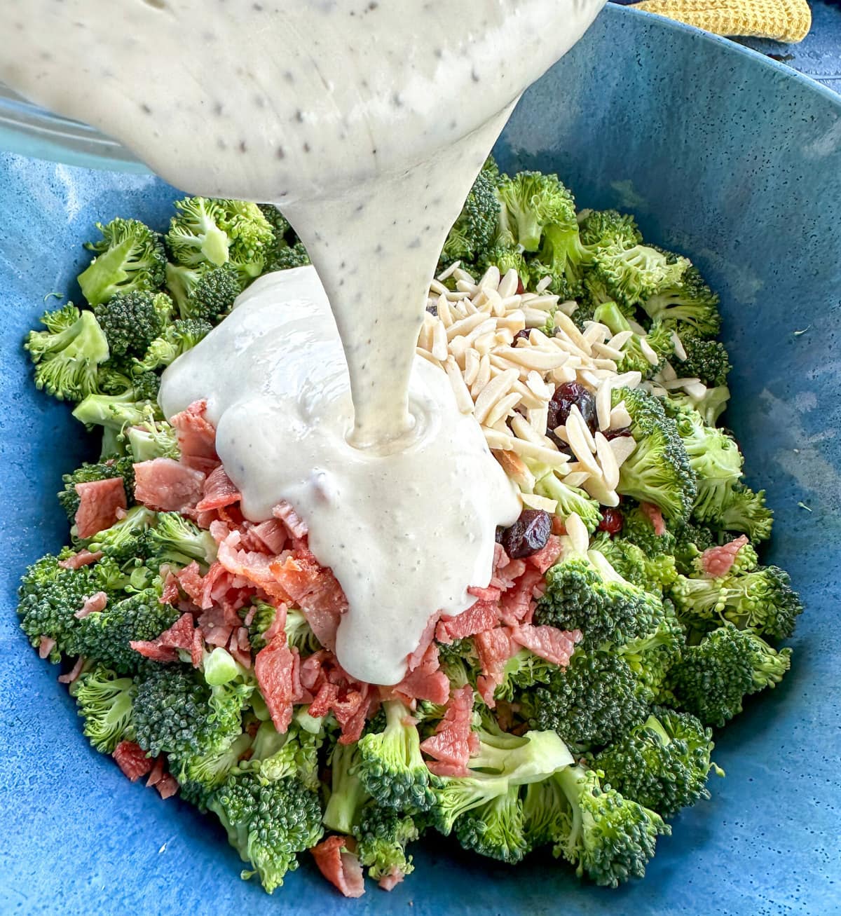 creamy coleslaw dressing being poured over bacon, cranberries, almonds and broccoli florets