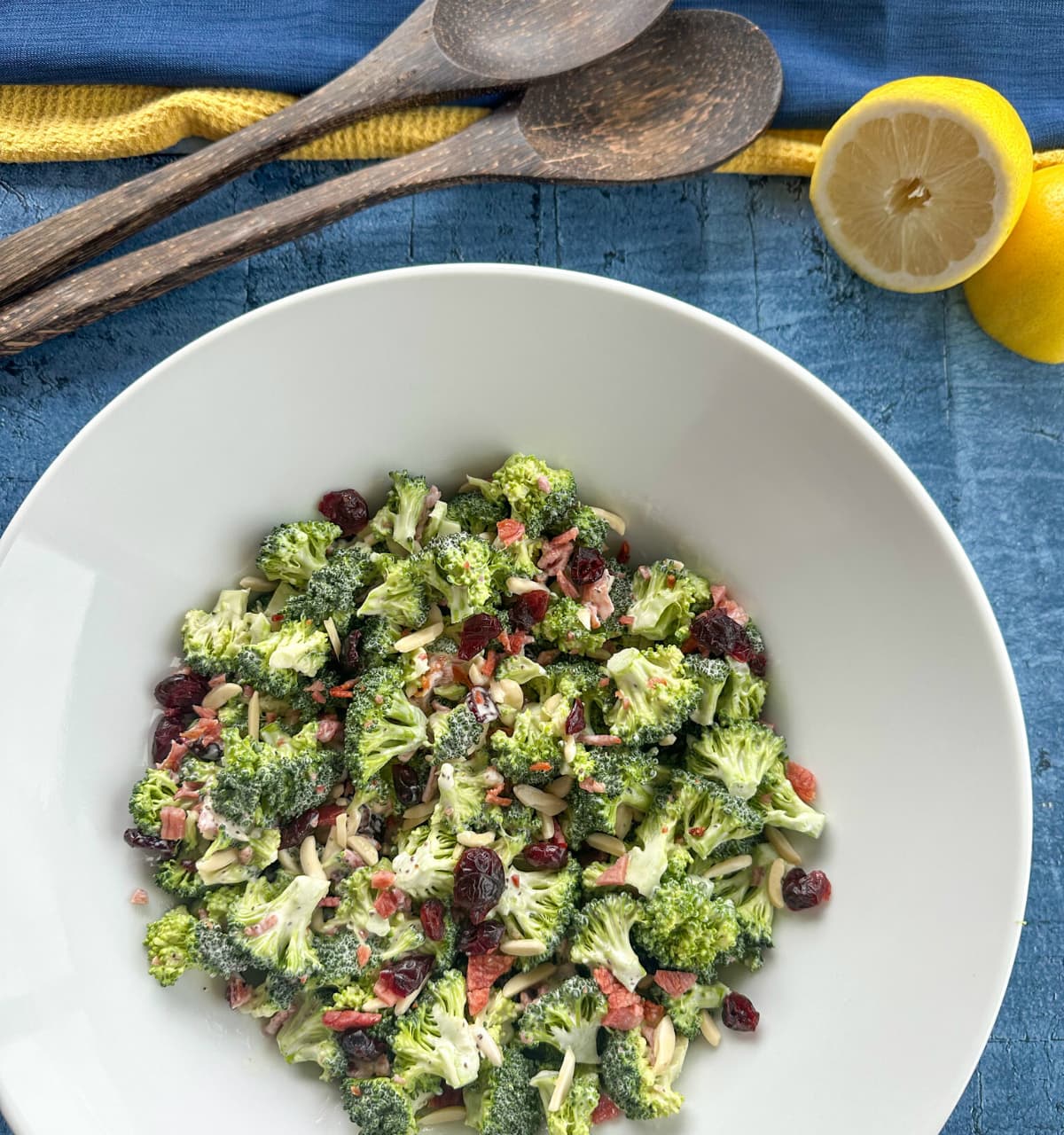 Creamy Broccoli Salad in a large white bowl with wooden salad serves and lemons