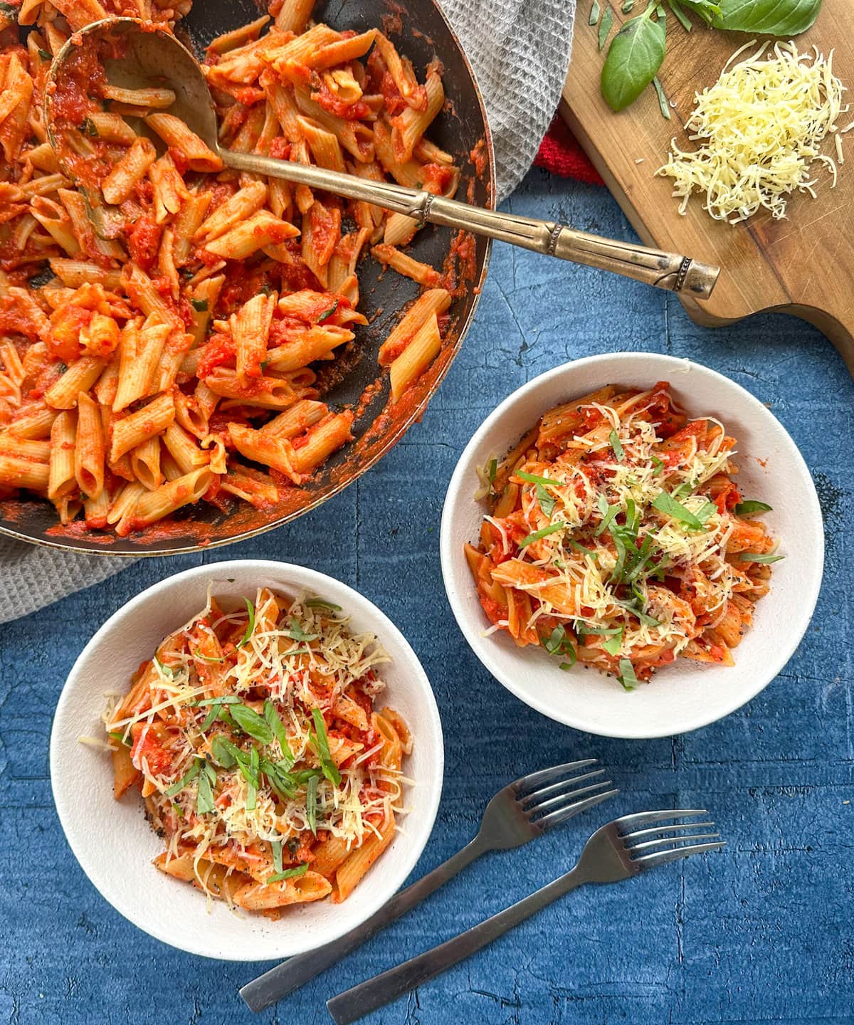Two white bowls with tomato and basil penne pasta with a frying pan full of pasta
