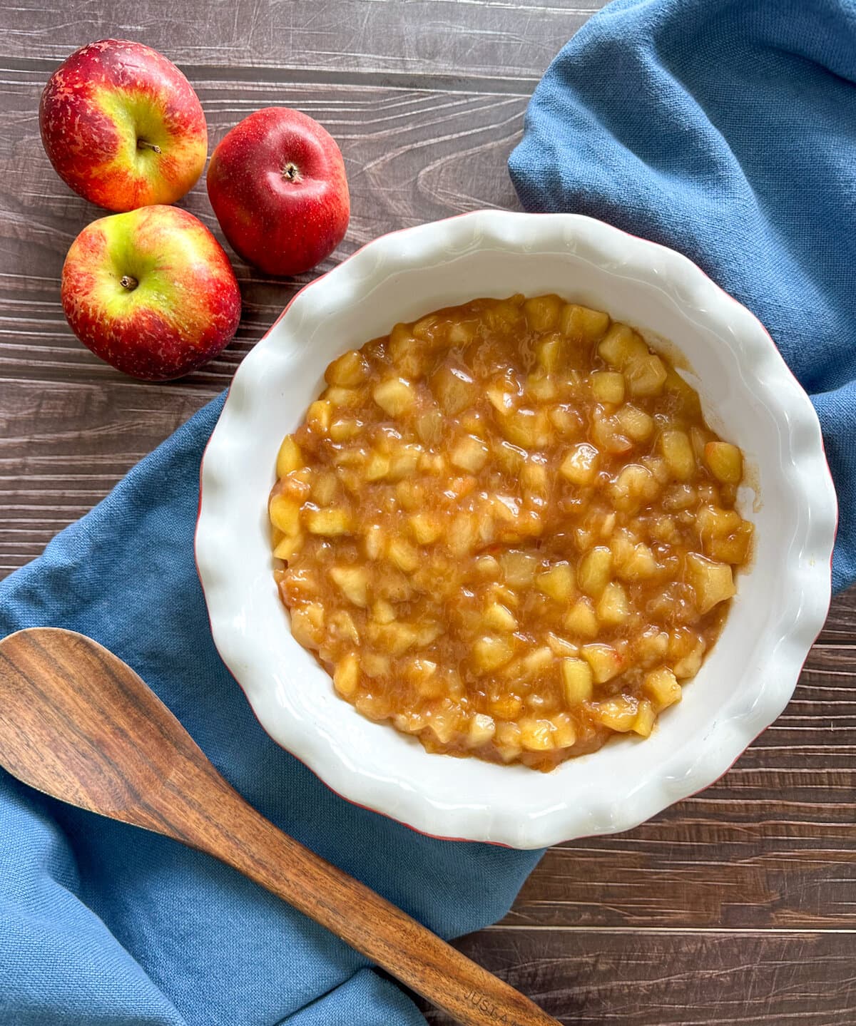 A pie dish with fresh homemade apple pie filling on a brown and blue background with fresh apples and a wooden spoon