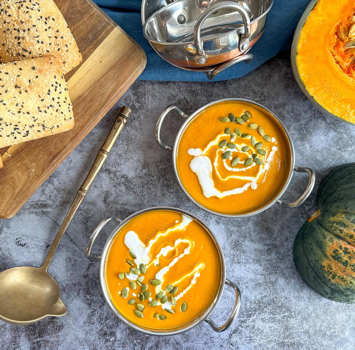 Two stainless steel bowls of Thai pumpkin soup set against a gray background with fresh pumpkin and bread rolls to the side. 
