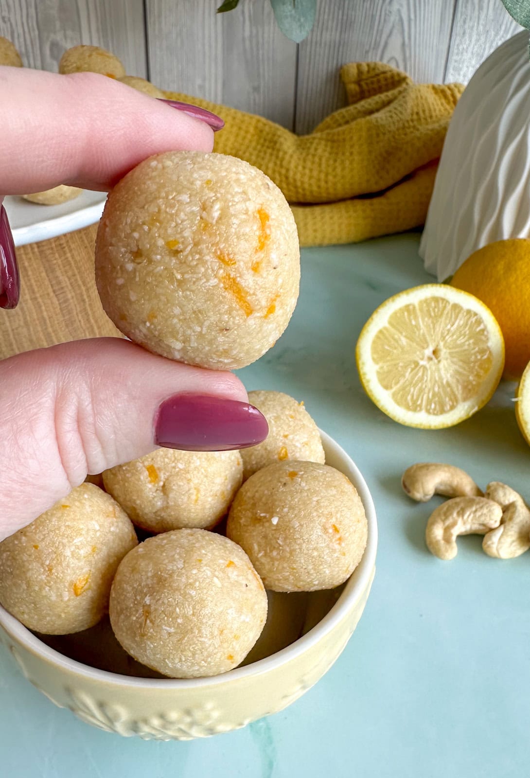 a lemon and cashew bliss ball being held between two fingers with fresh lemons 