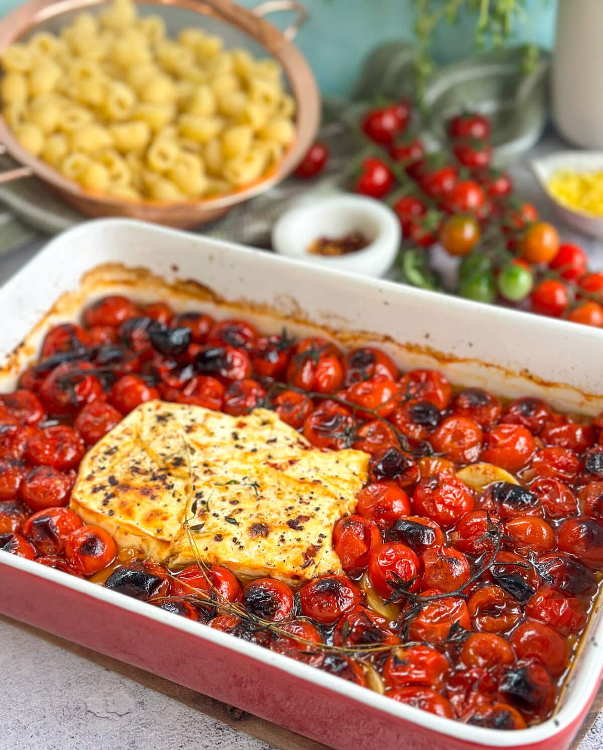 Large baking dish with golden brown feta cheese and charred burst cherry tomatoes with a selection of ingredients to add to finish the dish. 