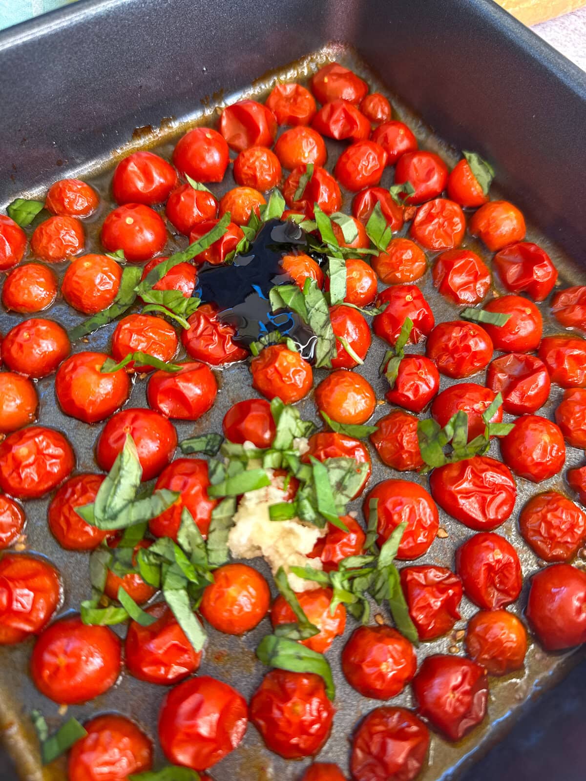 Adding balsamic glaze, garlic and basil to part roasted cherry tomatoes in a roasting dish